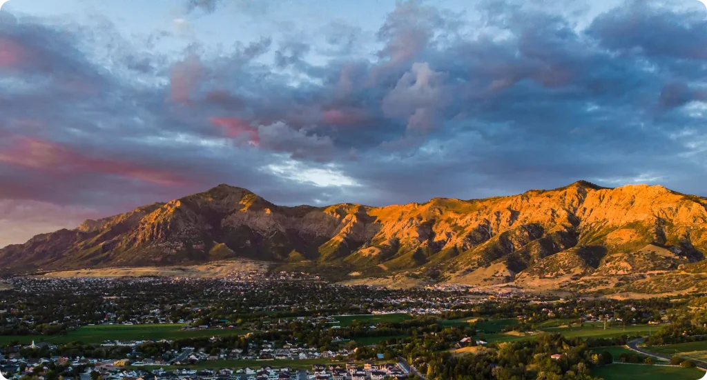 Utah neighborhood nestled below the Wasatch Mountains at sunset, with golden alpenglow on rugged peaks and pink clouds overhead.