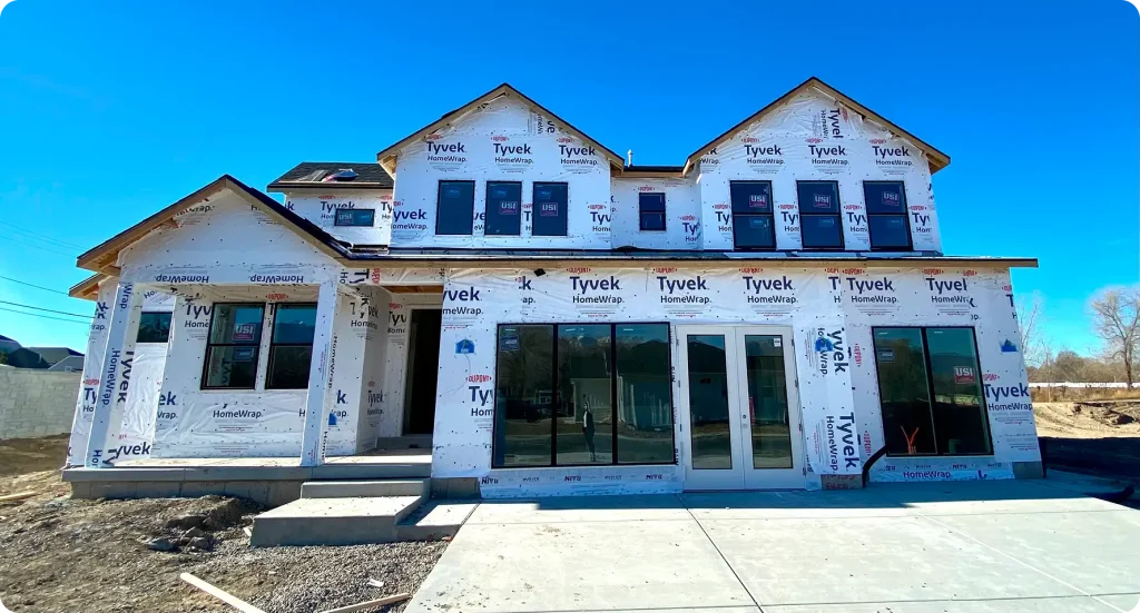 New home under construction in Utah wrapped in Tyvek HomeWrap with framed windows and concrete driveway under a clear blue sky.