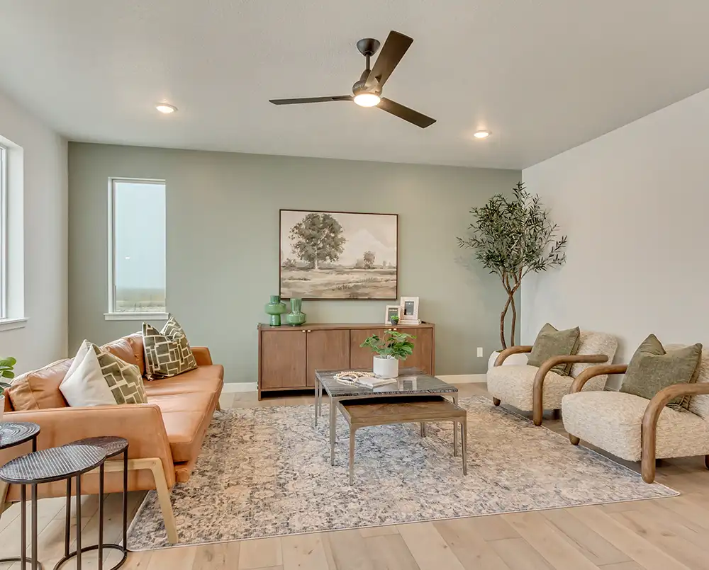 Living room with sage green accent wall, tan leather sofa, two armchairs, ceiling fan, landscape painting, and tall potted tree.