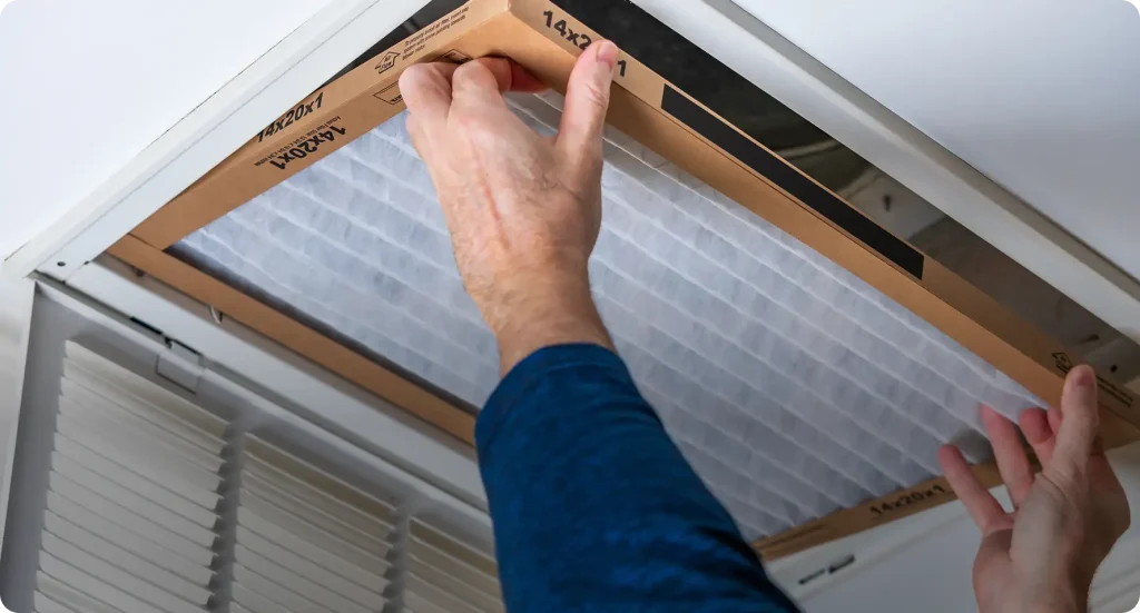 Homeowner sliding a fresh 14x20x1 pleated HVAC air filter into a ceiling return vent during routine new home maintenance.