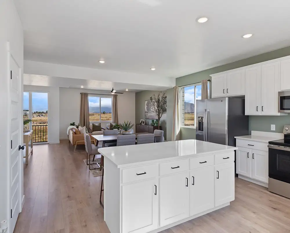 White kitchen island with stainless appliances opening to a dining table and living area with large windows and hardwood floors.