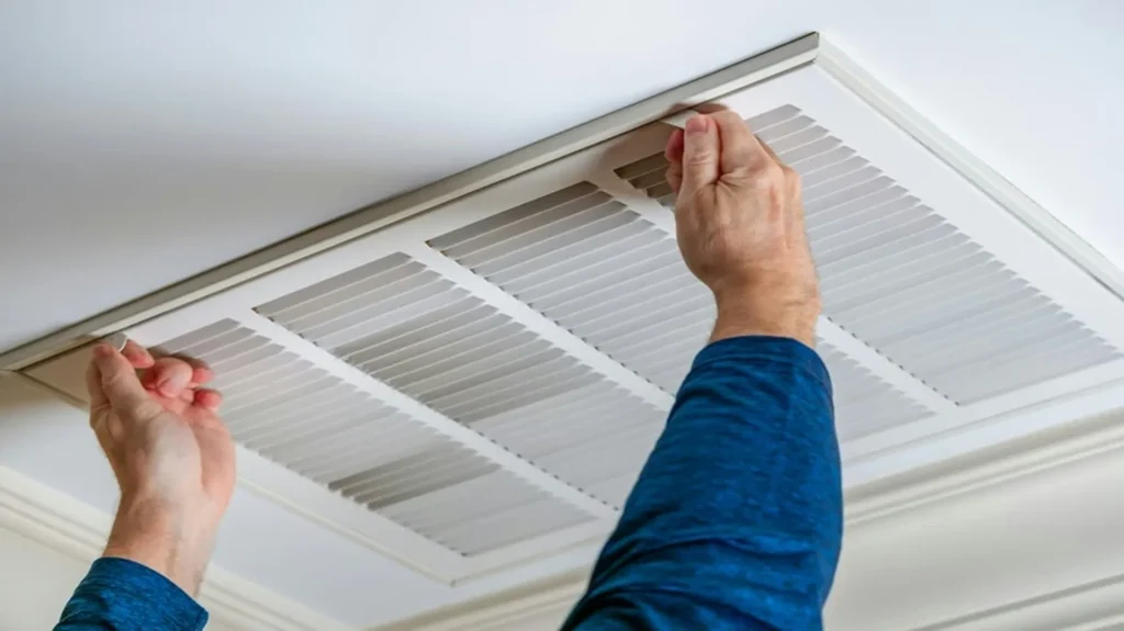 Person in blue shirt removing a white ceiling air return vent cover to access and replace the furnace filter inside.