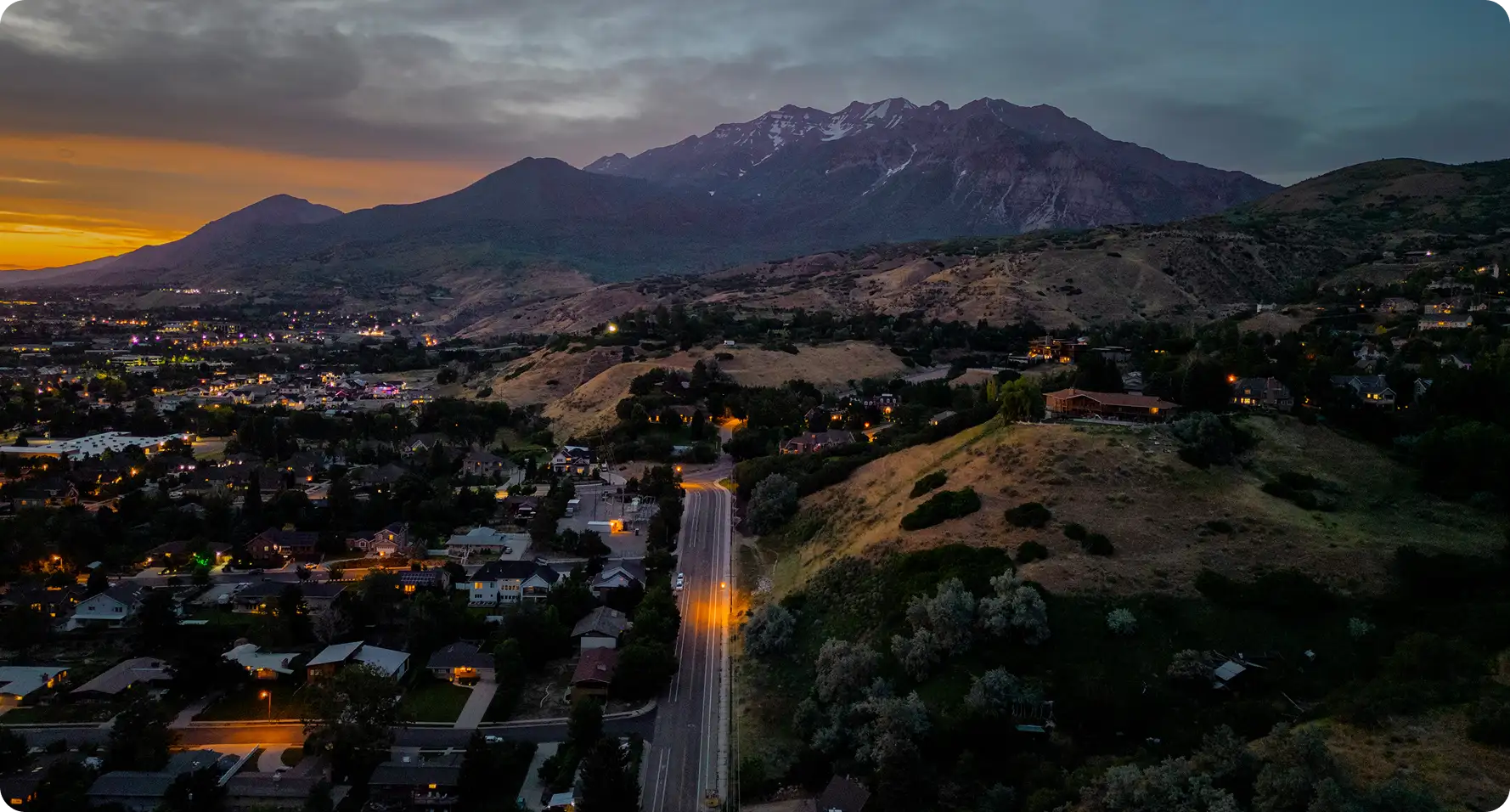Aerial dusk view of a Utah valley neighborhood with lit streets leading toward Mount Timpanogos beneath an orange sunset sky.