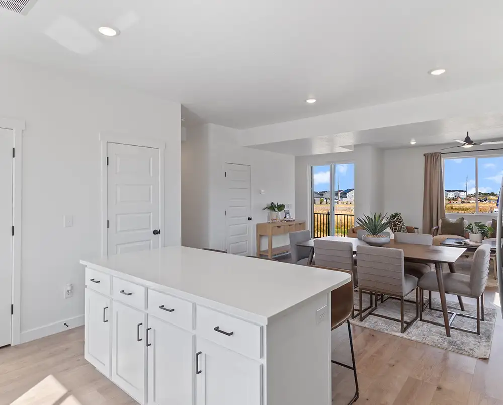 White kitchen island with shaker cabinets beside a wood dining table, upholstered chairs, and sliding glass doors to a backyard.