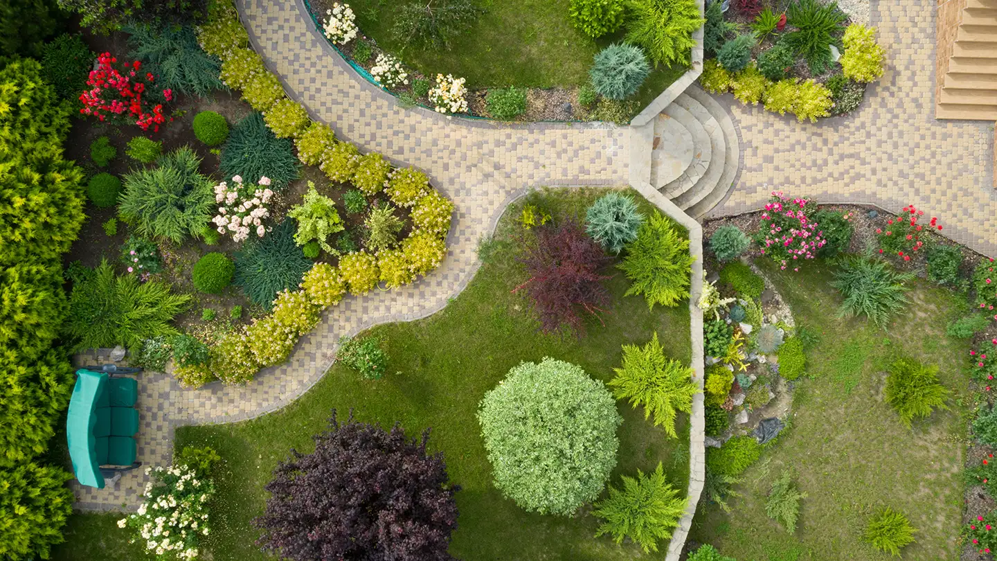 Overhead view of a landscaped backyard with curving stone paths, manicured shrubs, and colorful flower beds on a green lawn.