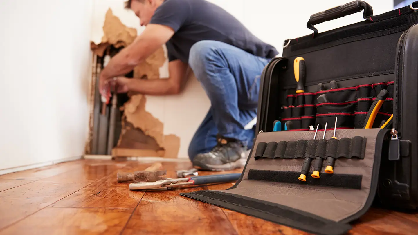 Man kneeling to repair plumbing behind a damaged drywall section, with an open tool bag and tools on a wood floor nearby.