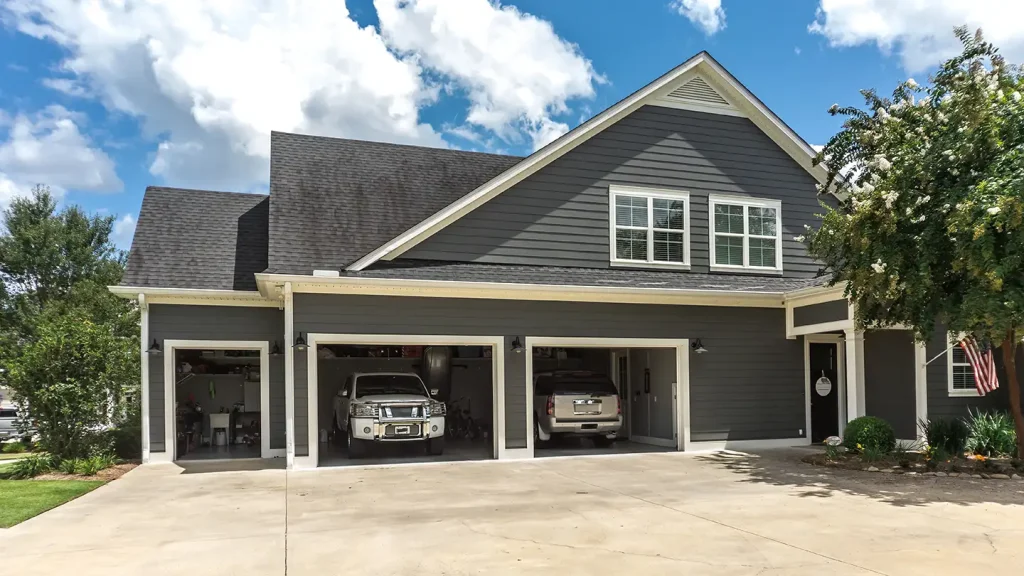 Gray two-story home with three open garage bays revealing two parked SUVs and a workshop under a partly cloudy blue sky.