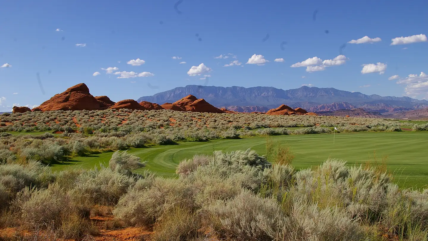 Manicured golf course green and fairway with desert sagebrush, red rock formations, and distant mountain ranges under blue sky.
