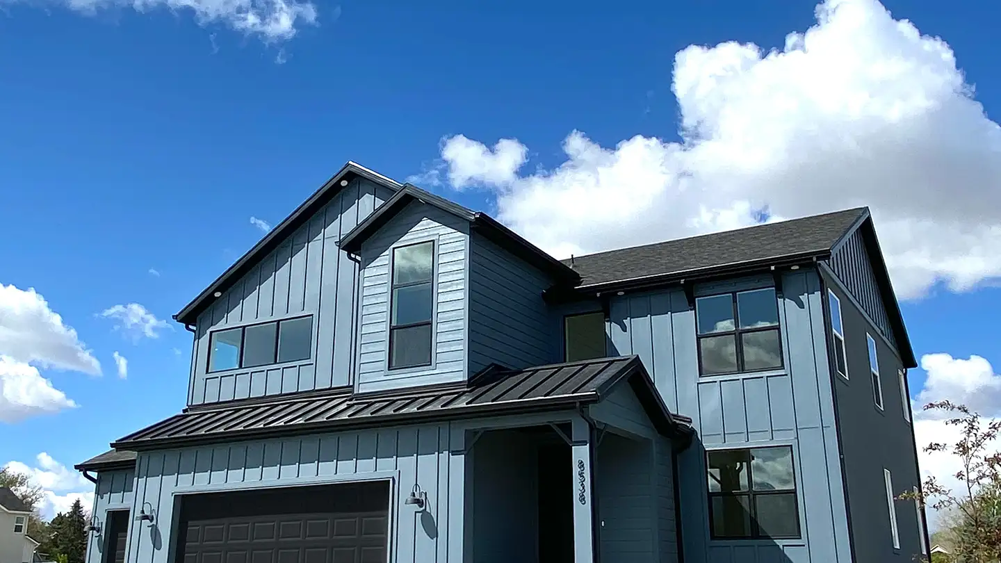 Modern two-story farmhouse with gray-blue board-and-batten siding and black metal porch roof against a cloud-filled blue sky.