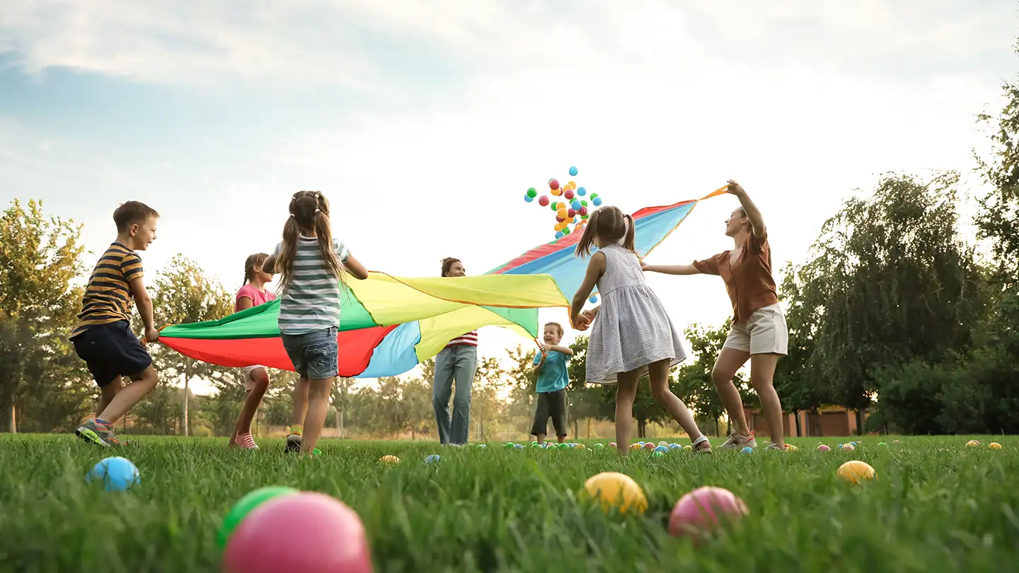 Children playing with a rainbow parachute in a grassy park, sending colorful plastic balls flying into the air on a sunny day.