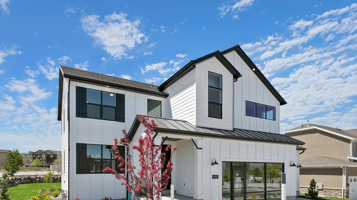 Two-story white modern farmhouse with black trim, a flowering pink tree, and a McArthur Homes sales sign under a blue sky.