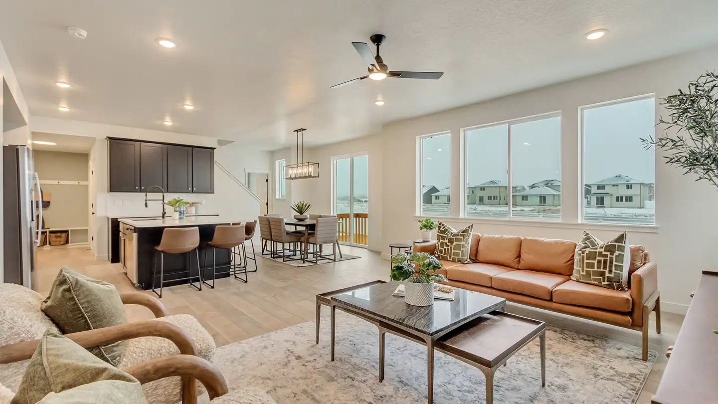Open-concept living room with a tan leather sofa, dining area, and kitchen island with bar stools beneath bright windows.