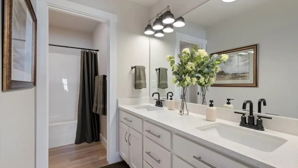 Modern bathroom with a white double-sink vanity, black fixtures, framed mirror, and white hydrangeas beside a tub-shower combo.