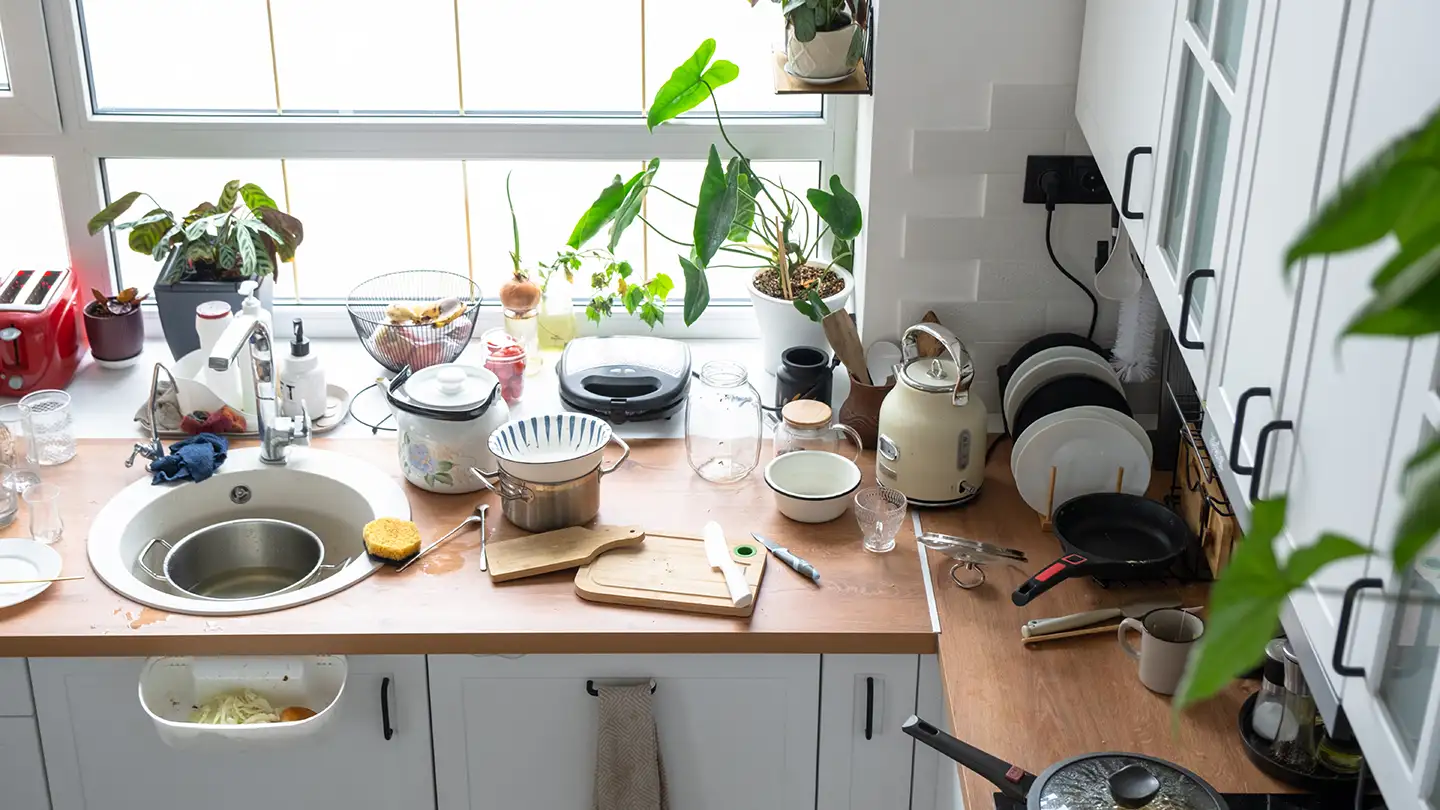 Cluttered kitchen counter overflowing with dishes, pots, a kettle, and houseplants on the windowsill in a small kitchen.