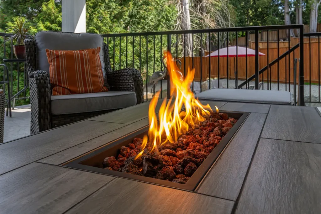Flames rise from lava rocks in a sleek rectangular DIY fire pit on a covered backyard deck beside a wicker patio chair.