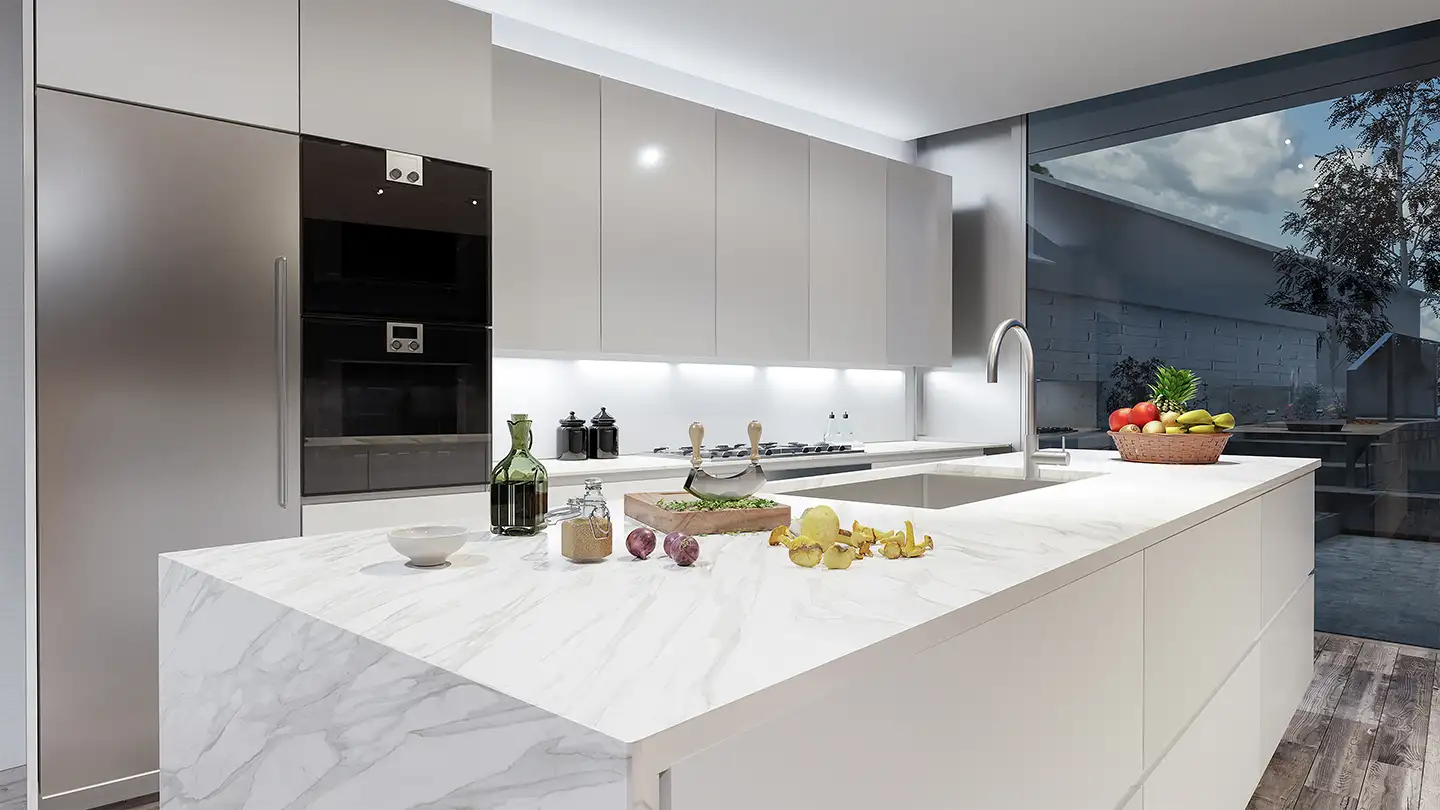 Modern kitchen with a white marble island, light gray cabinets, built-in black ovens, and a fruit basket by large windows.