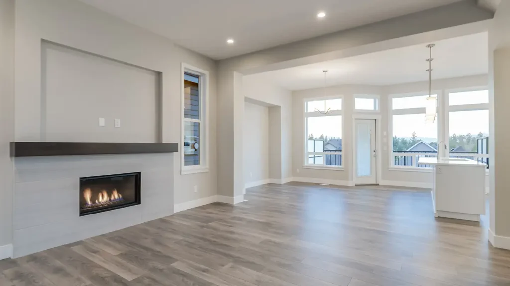 Empty living and dining area with a lit linear fireplace, hardwood floors, large windows, and a chandelier overhead.