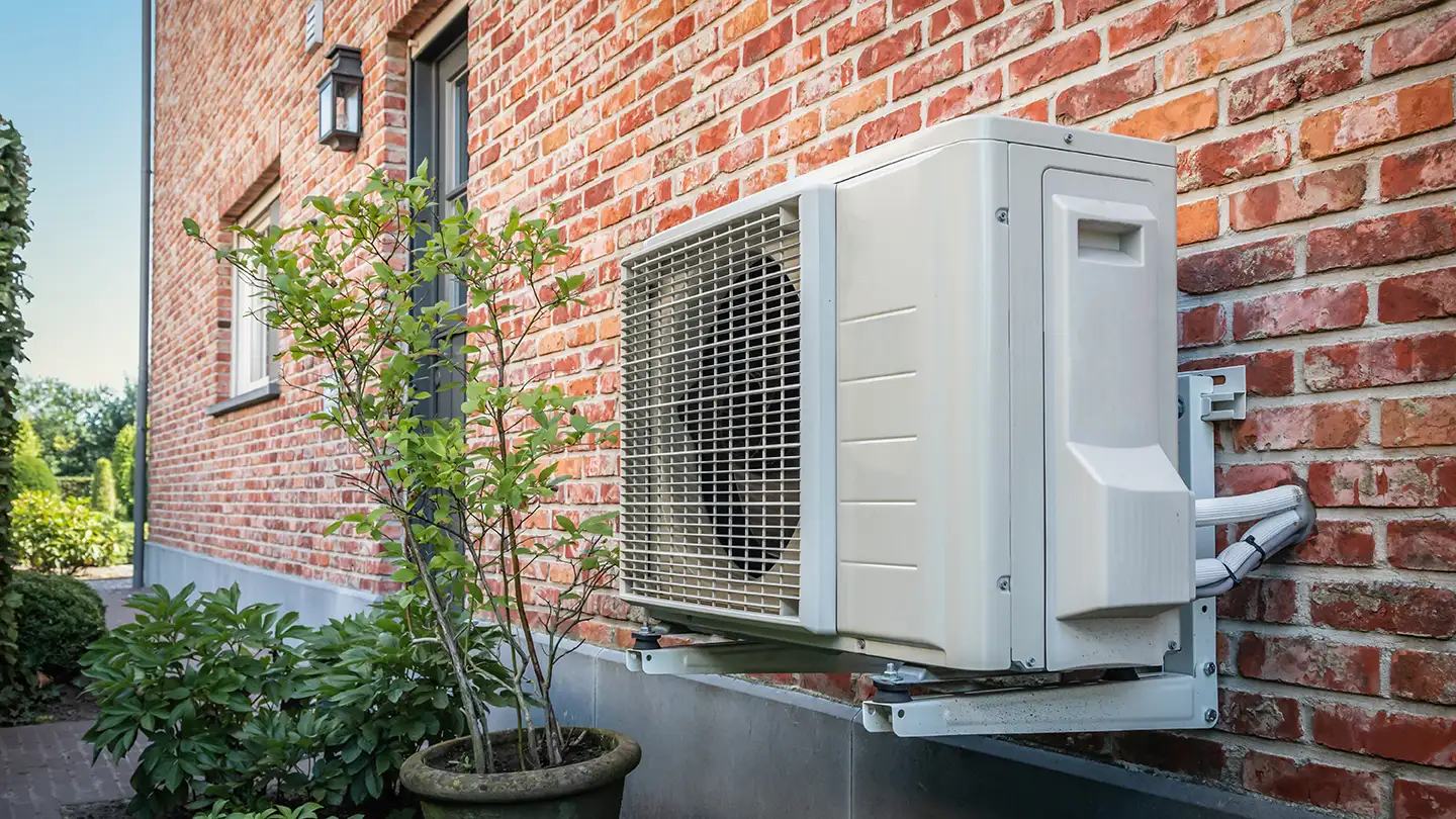 White outdoor air conditioning unit mounted on the red brick exterior of a home, with green shrubs beside it.