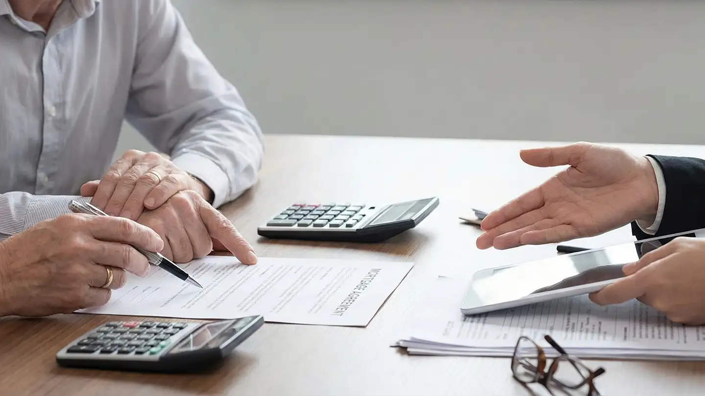 Two people at a desk reviewing a mortgage agreement with calculators, a tablet, pen, and reading glasses on the surface.