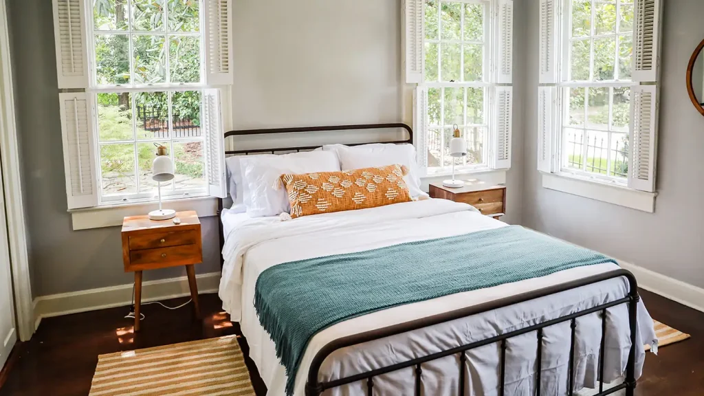 Cozy guest bedroom with a black iron bed, white bedding, teal throw, orange lumbar pillow, and tall windows with shutters.