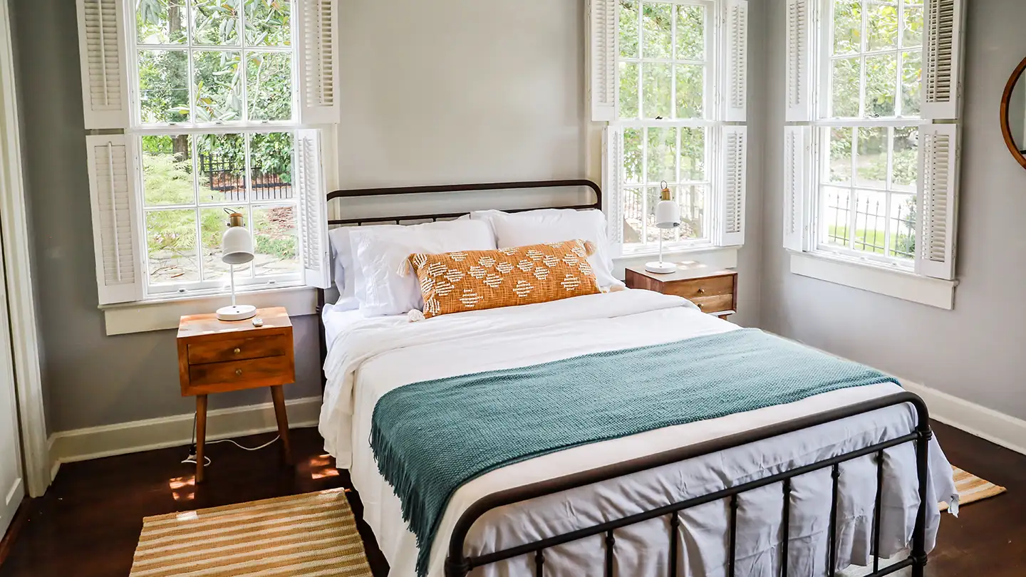 Cozy guest bedroom with a black iron bed, white bedding, teal throw, orange lumbar pillow, and tall windows with shutters.