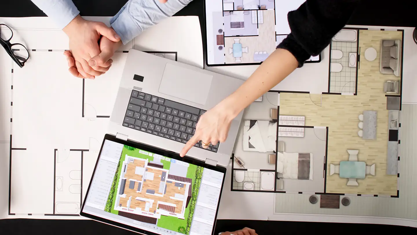 Overhead view of two people reviewing a laptop with 3D home design software, surrounded by printed and colored floor plans.