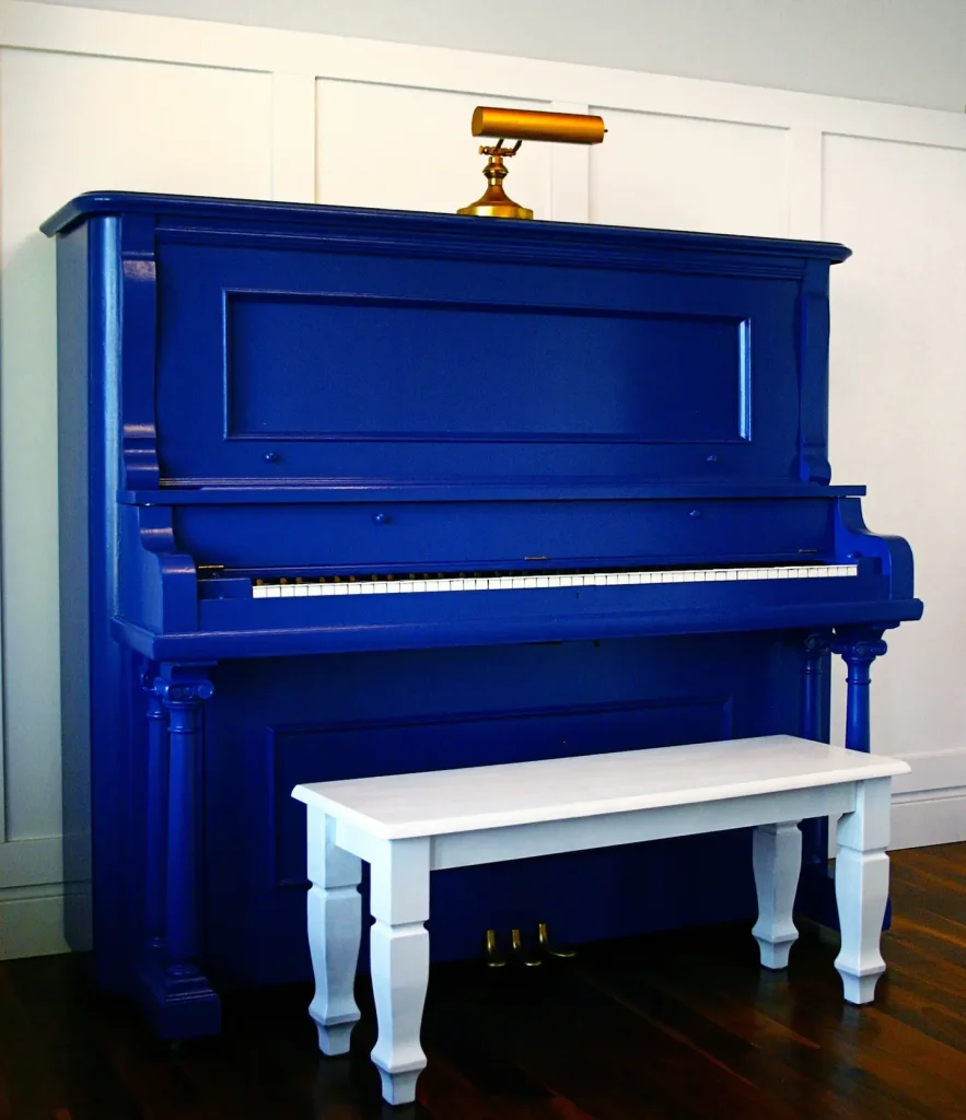 Refinished royal blue upright piano with a brass piano light and white wooden bench on dark hardwood floor against a board-and-batten wall.