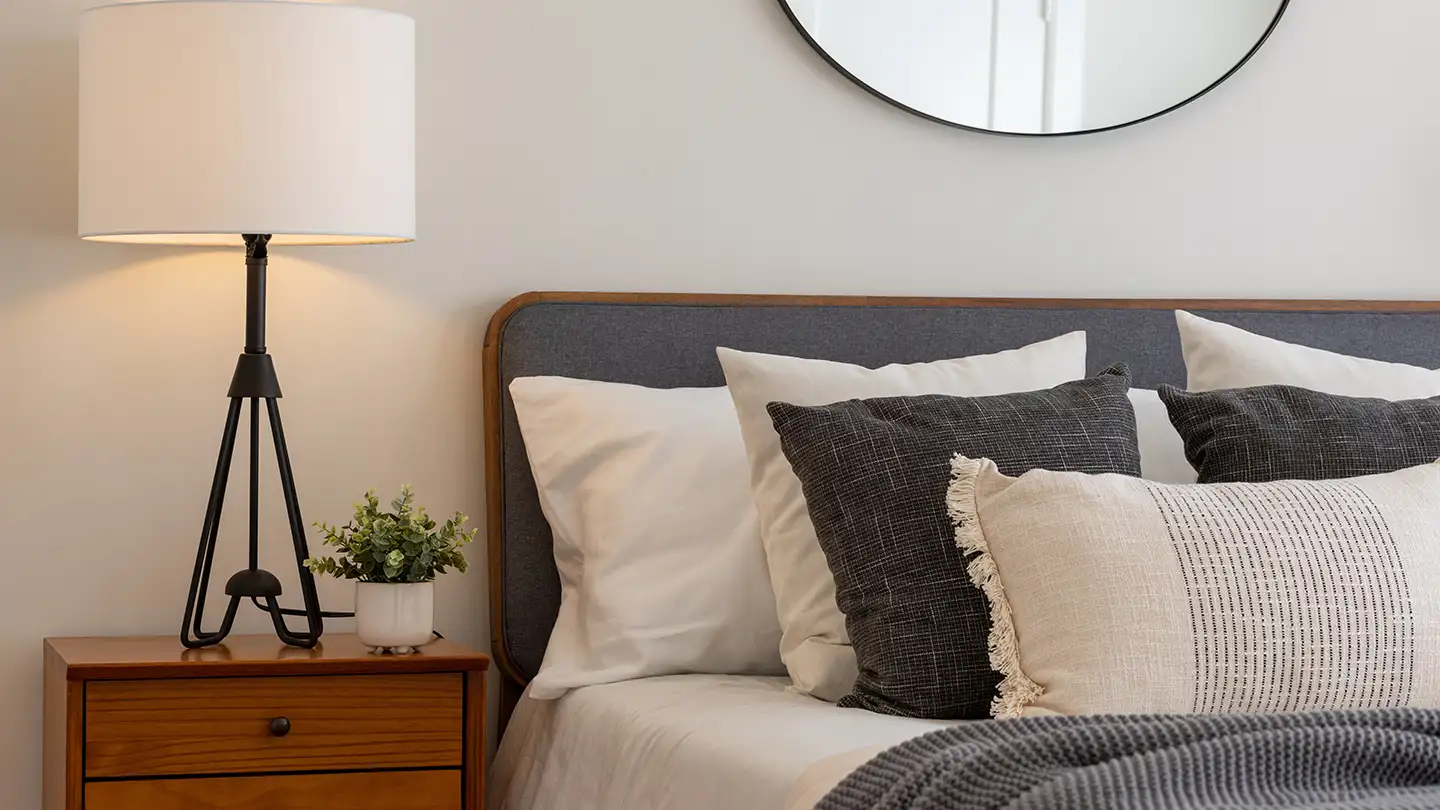 Bedroom corner with an upholstered gray headboard, layered pillows, a tripod lamp on a wood nightstand, and a round mirror.