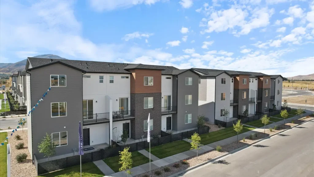 Row of three-story modern townhomes with mixed gray, white, and wood siding lined by young trees with mountains behind.