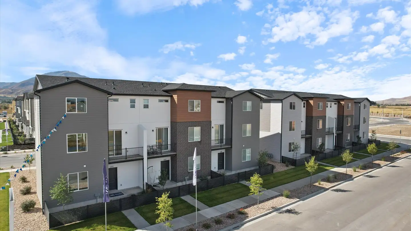 Row of three-story modern townhomes with mixed gray, white, and wood siding lined by young trees with mountains behind.