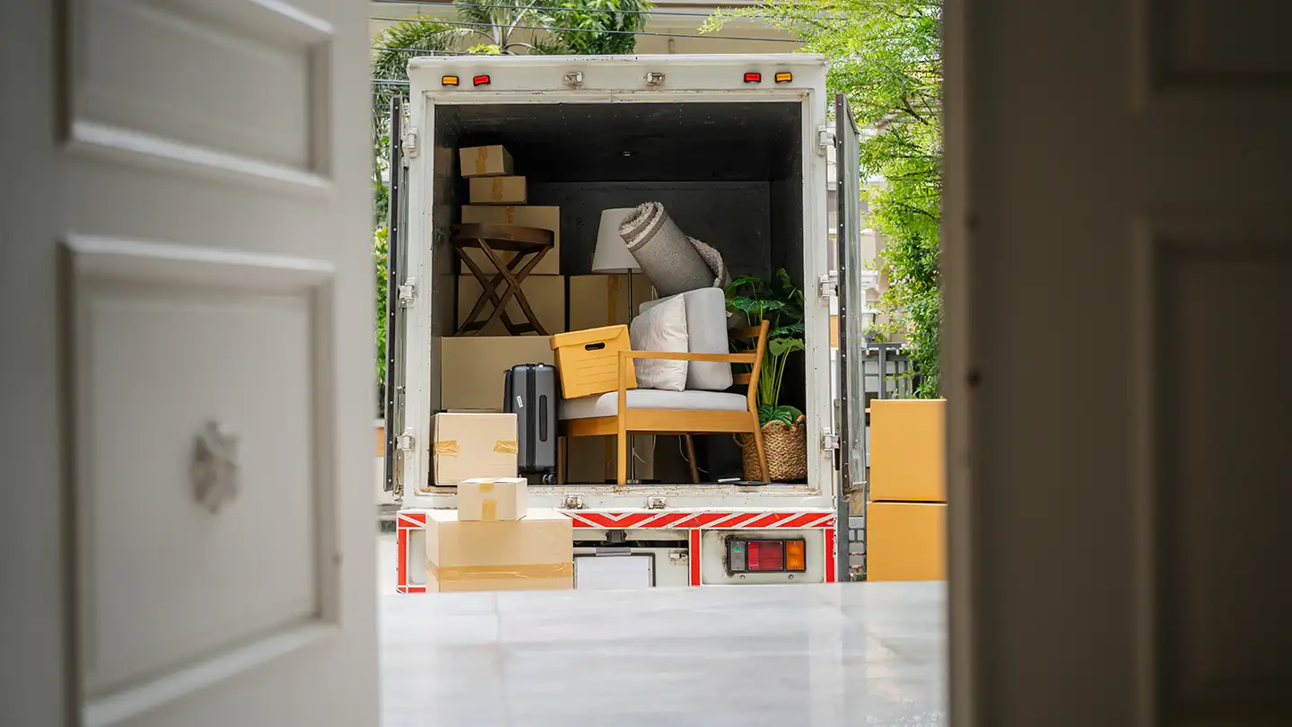 View through an open doorway of a moving truck packed with cardboard boxes, a chair, suitcase, lamp, and houseplants.