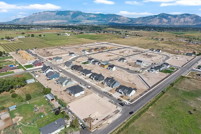 Aerial view of a new residential subdivision under construction with framed houses, dirt lots, and farmland set against a mountain range.