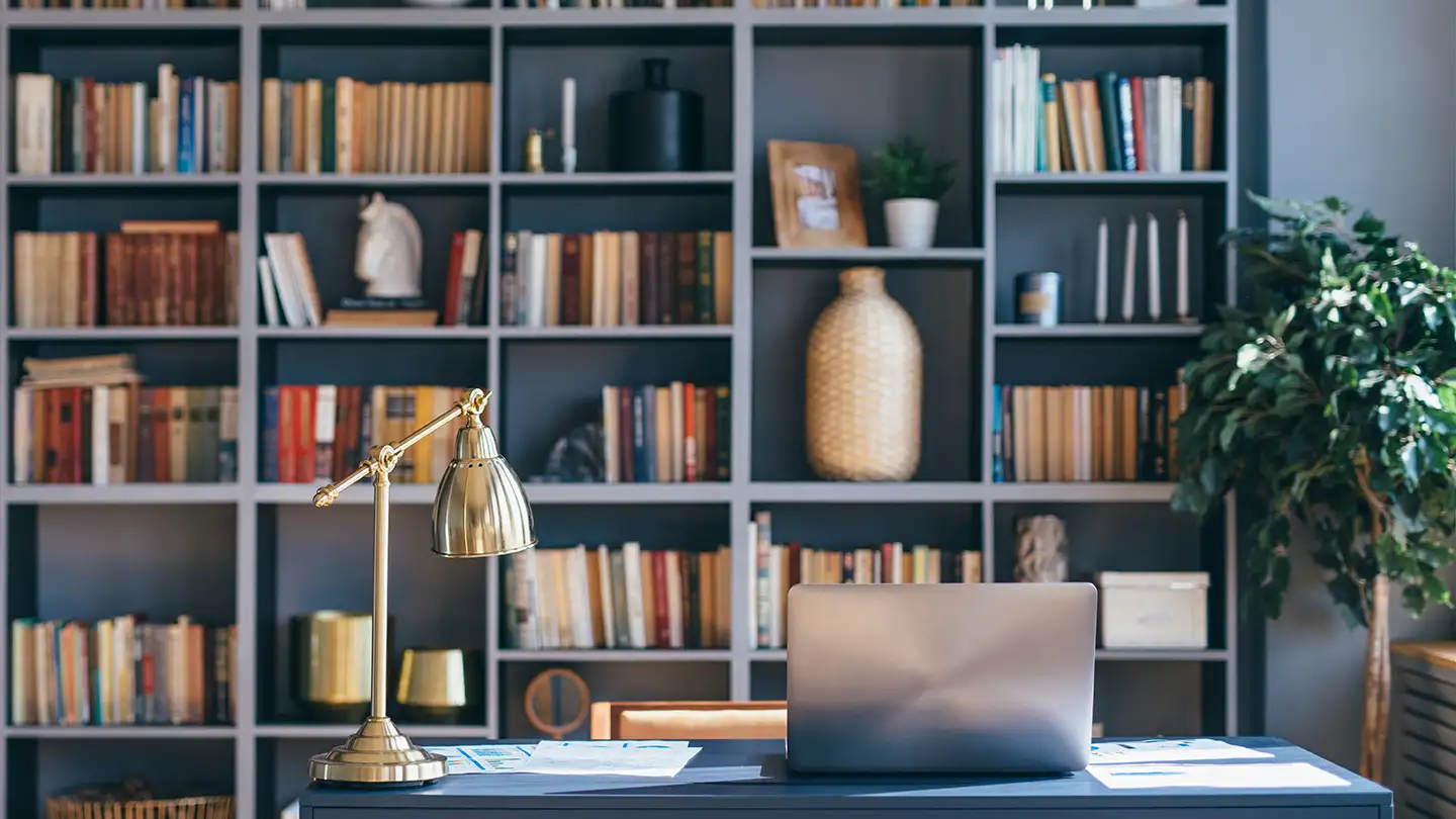 Home library with tall dark built-in bookshelves filled with books and decor, a brass desk lamp, and a closed laptop on a desk.