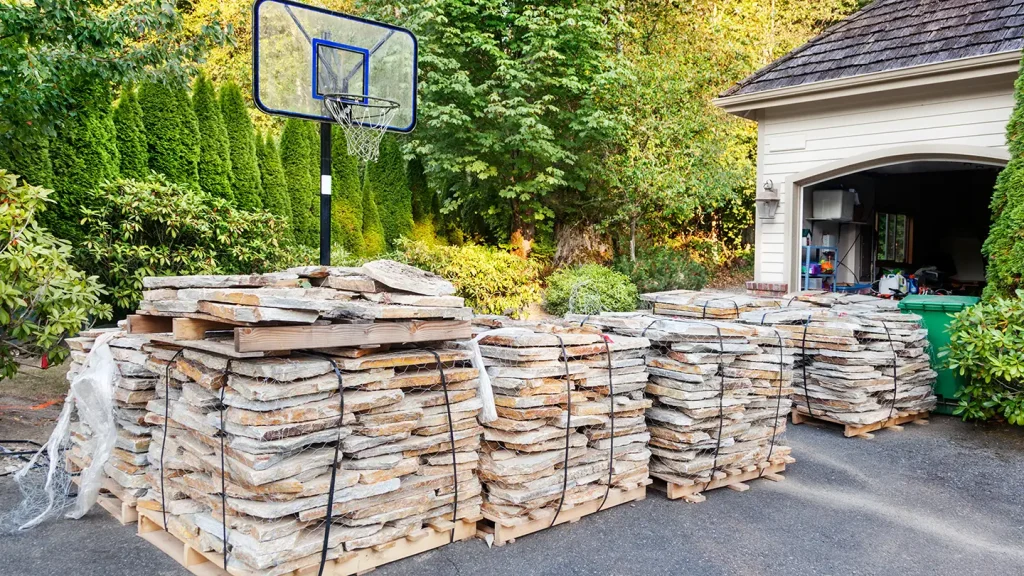 Pallets of stacked stone veneer in a driveway beside a basketball hoop and an open garage at a home.