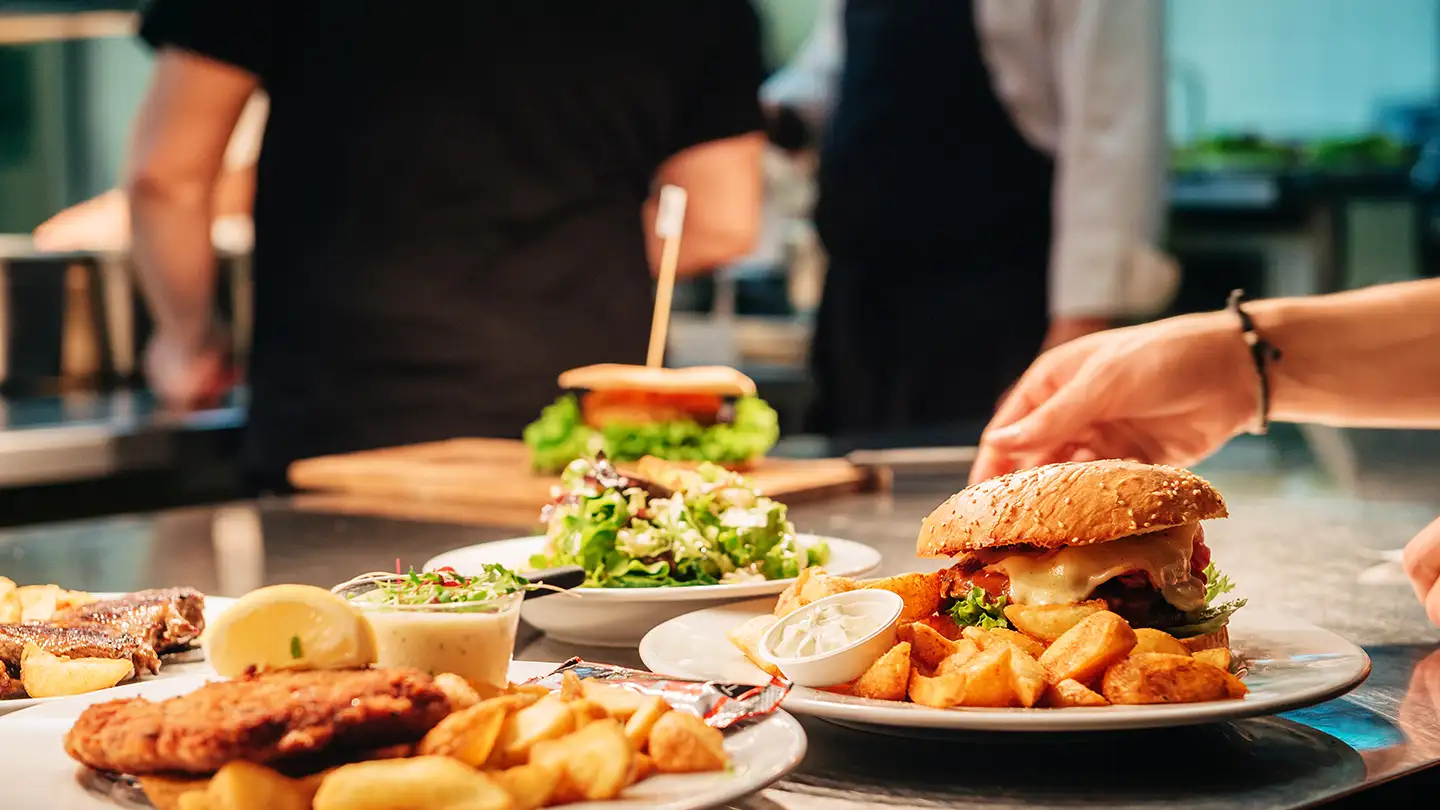 Plates of cheeseburgers, fries, salad, and fish being prepared and served by chefs in a busy restaurant kitchen.