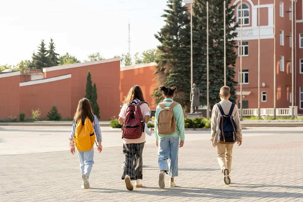 Four students with colorful backpacks walk together toward a red brick school building on a sunlit campus walkway.
