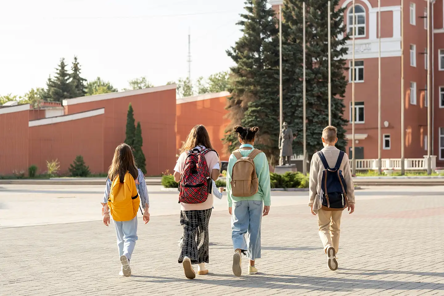 Four students with colorful backpacks walk together toward a red brick school building on a sunlit campus walkway.