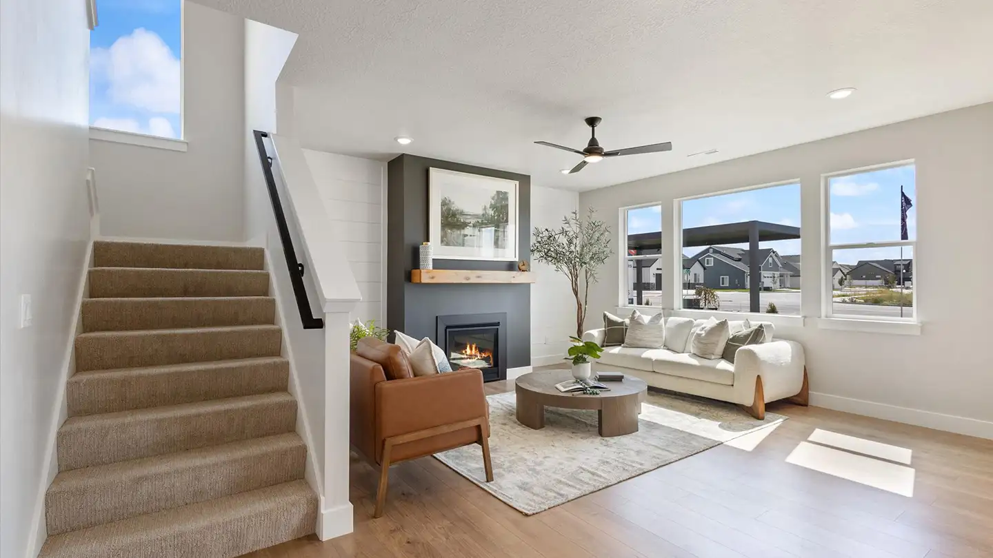 Bright living room with a carpeted staircase, lit gas fireplace, white sofa, brown leather chair, and a round coffee table.
