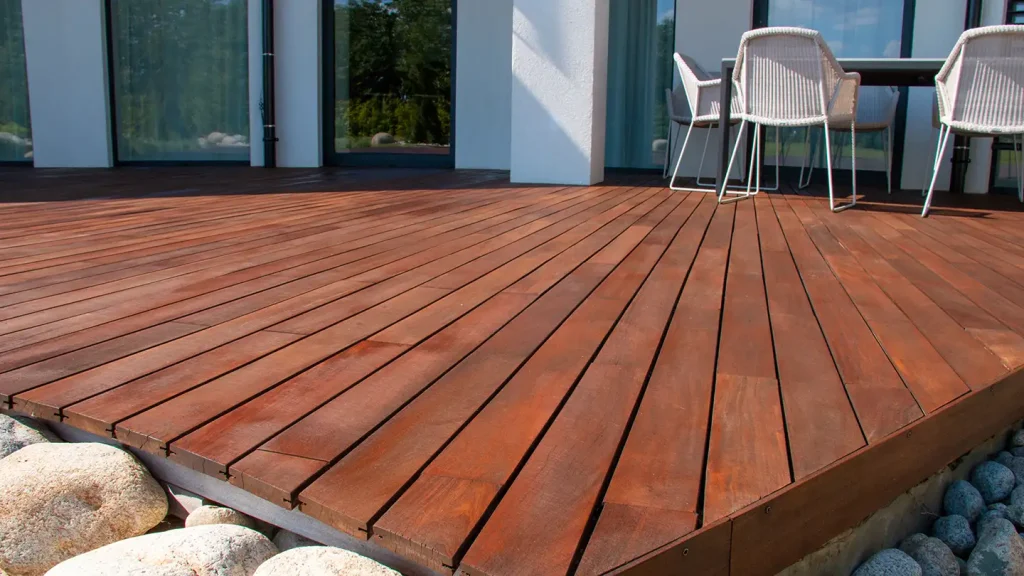 Stained wood deck with white wicker chairs near sliding glass doors and a border of smooth river rocks at its edge.