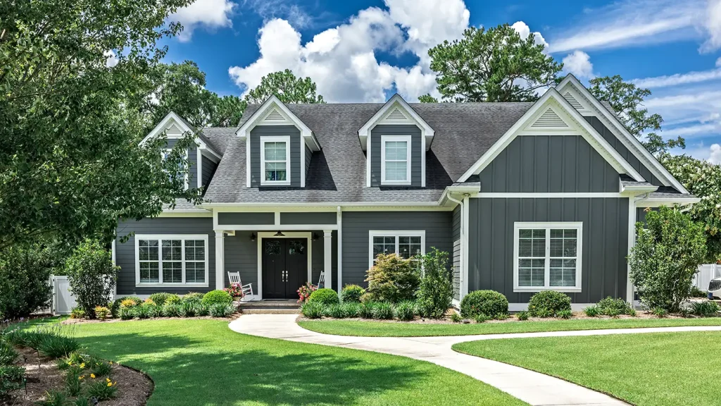 Dark gray craftsman home with white trim, lush landscaped shrubs, a green lawn, and a curving walkway under a partly cloudy sky.