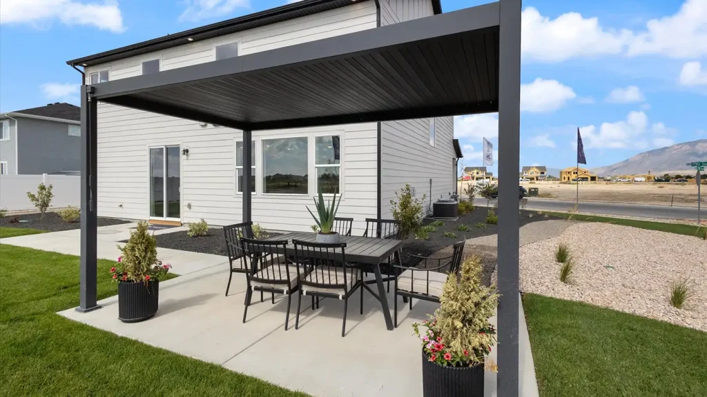 Backyard patio with a black metal pergola, dining table for six, potted flowers, and a white McArthur home behind.
