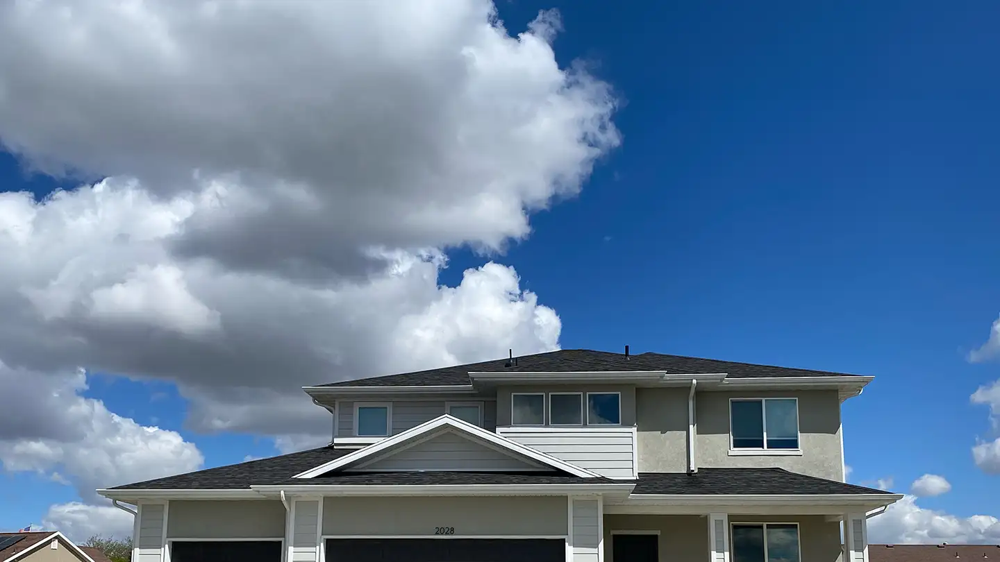 Two-story tan and gray home exterior framed by large fluffy white cumulus clouds against a deep blue sky.
