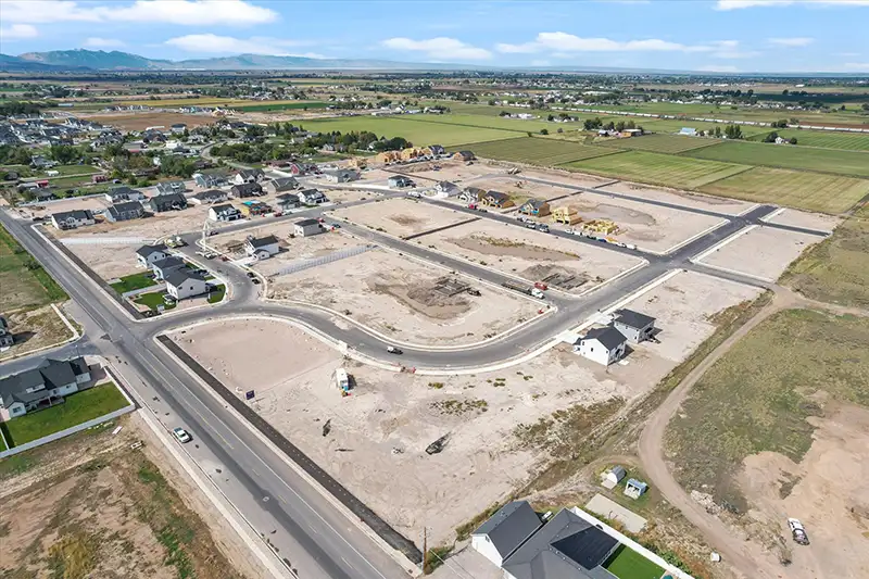 Aerial view of a new housing development with empty graded lots, curving paved streets, and surrounding farmland under scattered clouds.