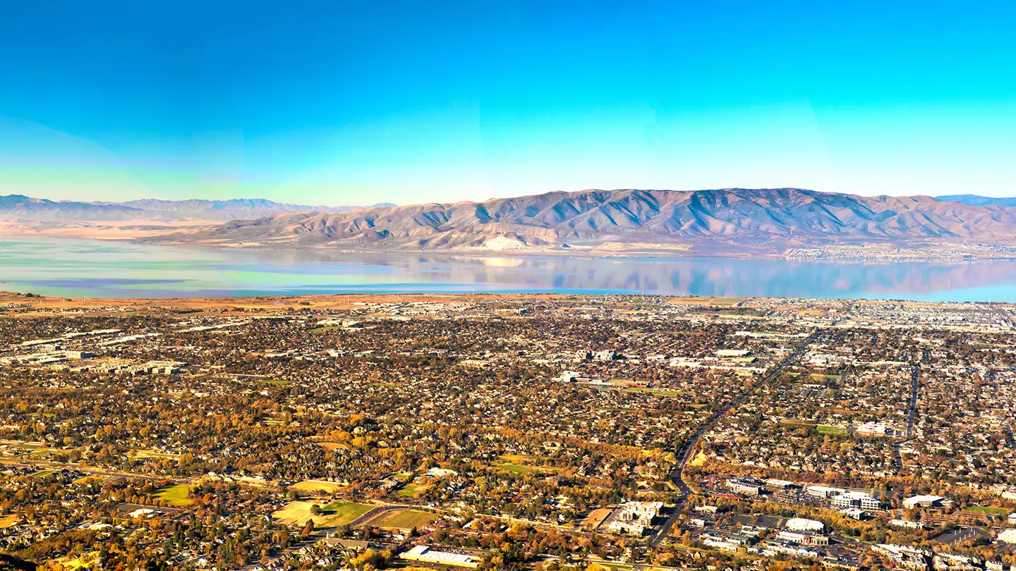 Aerial view of a Utah valley city with sprawling neighborhoods extending toward Utah Lake and a mountain range on the horizon.
