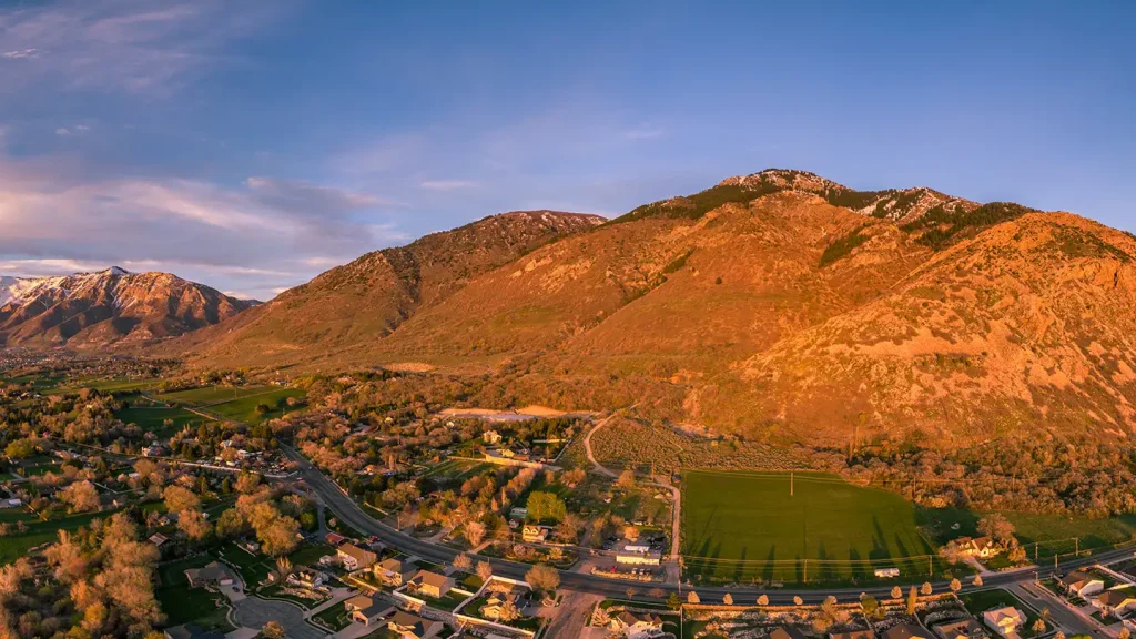 Aerial sunset view of an Ogden neighborhood backed by glowing golden mountains, with a soccer field and homes in the foreground.