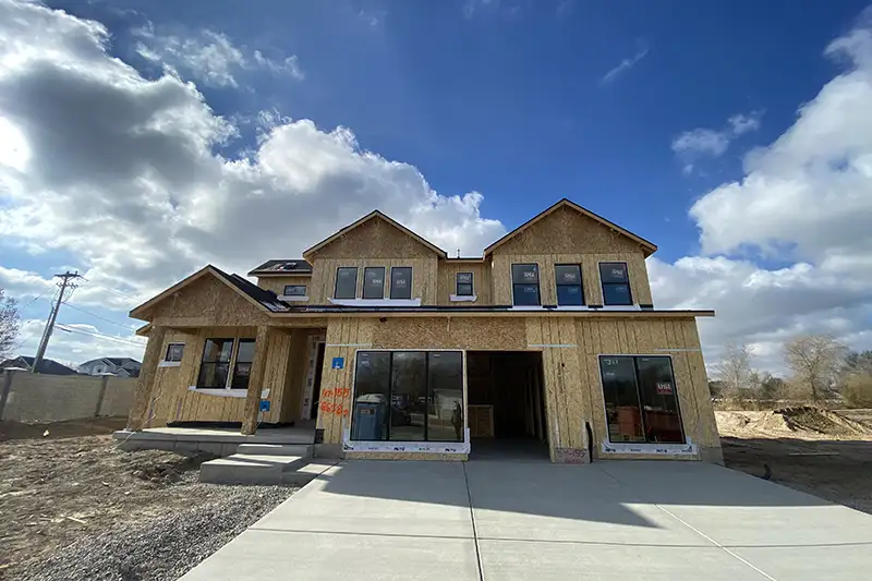 Two-story home under construction with exposed OSB sheathing, dormer windows, and a covered porch on a concrete driveway under a partly cloudy sky.