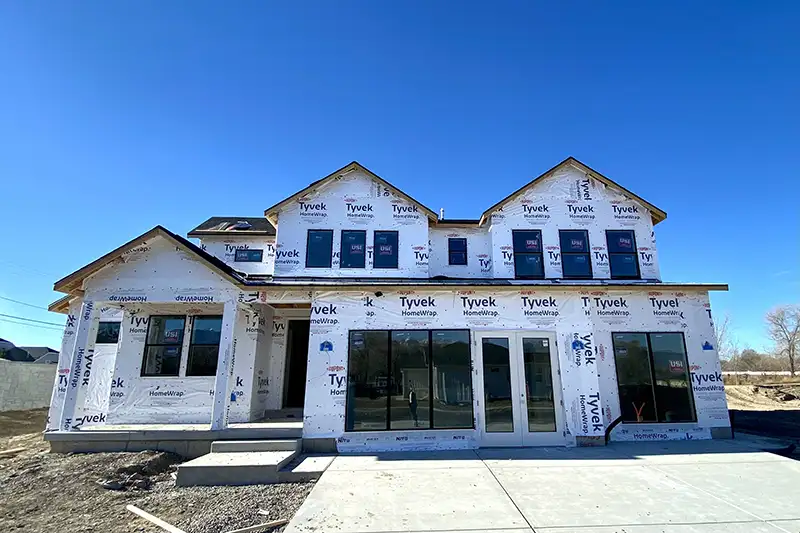Two-story home in the framing stage wrapped in Tyvek HomeWrap, with dormers and large window openings under a clear blue sky.