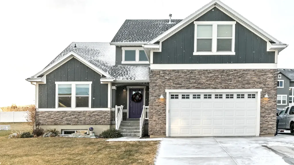 Dark gray craftsman home with stone accents, white trim, a two-car garage, snow on the roof, and a wreath on the front door.