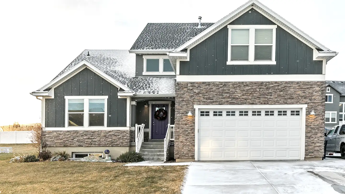 Dark gray craftsman home with stone accents, white trim, a two-car garage, snow on the roof, and a wreath on the front door.