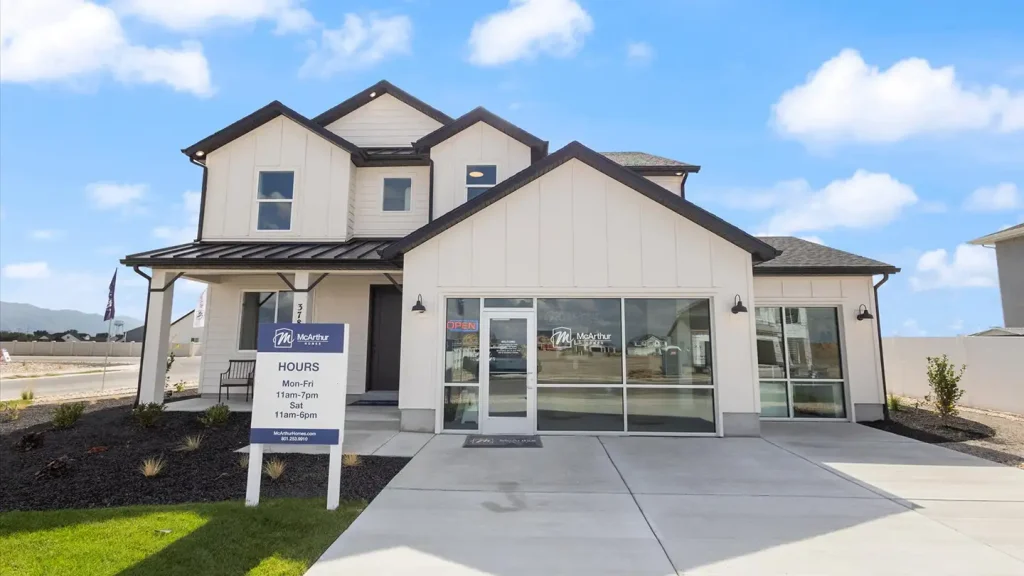 White two-story McArthur Homes model home with attached sales office storefront and a sign listing open hours below a blue sky.